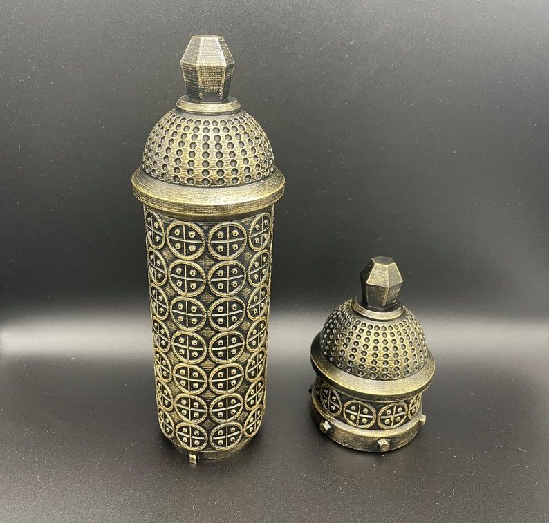 May include: Two gold-colored decorative containers with intricate patterns. The larger container is taller and has a more ornate design. The smaller container is shorter and has a simpler design.
