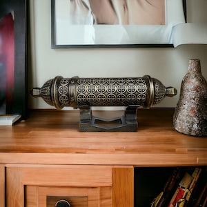 May include: A decorative cannon shaped box with a black and gold finish. The box has a detailed design of circles and is sitting on a wooden surface.