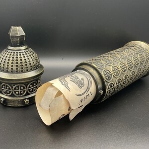 May include: A gold-colored metal container with a scroll inside. The container has a decorative design of circles and squares. The scroll is unfurled and has a hand-drawn design on it. The text on the scroll reads "...LYM1".
