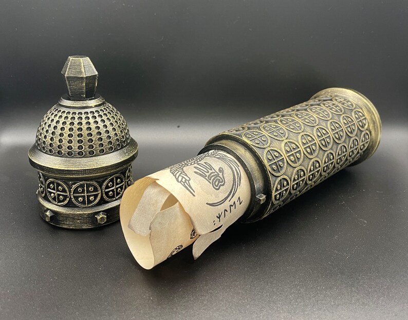 May include: A gold-colored metal container with a scroll inside. The container has a decorative design of circles and squares. The scroll is unfurled and has a hand-drawn design on it. The text on the scroll reads "...LYM1".