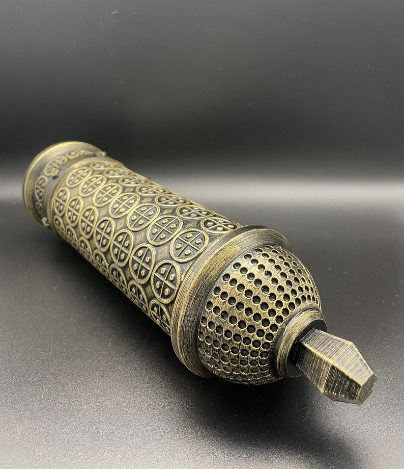 May include: A gold and black decorative cylinder with a patterned design. The cylinder has a small, pointed tip at one end.