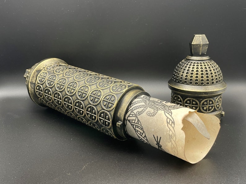May include: A decorative, ornate, gold-colored metal box with a scroll inside. The box has a detailed design of repeating circles and a domed lid.