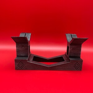 May include: A black plastic stand with a geometric design. The stand is designed to hold two items, such as video game controllers.