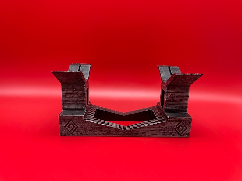 May include: A black plastic stand with a geometric design. The stand is designed to hold two items, such as video game controllers.