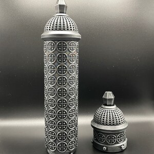 May include: Two silver metal containers with intricate designs. The larger container is taller and has a domed top with a circular pattern. The smaller container is shorter and has a similar domed top with a circular pattern.