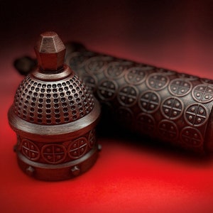 May include: A close-up of a decorative metal bottle stopper with a domed top and intricate details. The stopper is made of a dark metal with a textured finish. The stopper is lying on a red surface.