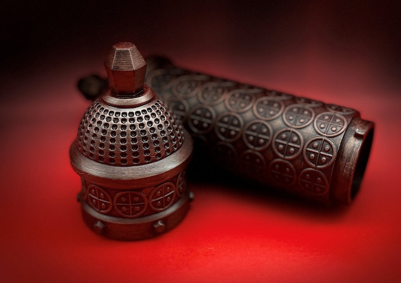 May include: A close-up of a decorative metal bottle stopper with a domed top and intricate details. The stopper is made of a dark metal with a textured finish. The stopper is lying on a red surface.