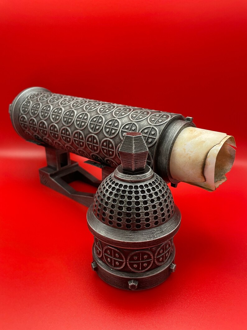 May include: A silver metal decorative box with a scroll inside. The box has a detailed design of circles and crosses. The box is on a red surface.