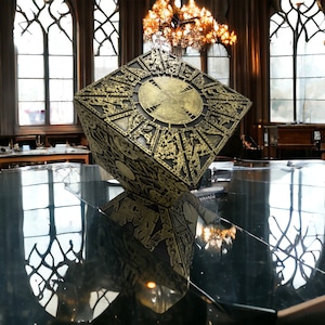 May include: An intricate, cube-shaped puzzle box with a circular design on the top, finished in gold. The box is resting on a glass surface, reflecting the surrounding room.
