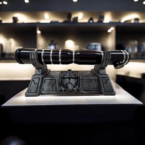 May include: A black lightsaber with silver detailing is displayed on a grey stand, featuring a helmet design. The lightsaber is cylindrical with silver bands. The stand has a rectangular base and is on a white surface. The item is displayed indoors.