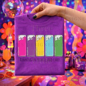 May include: Purple t-shirt featuring colorful Red Bull can graphics in pink, purple, yellow, blue, and green. The text "RUNNING ON REDBULL AND CHAOS" is printed below the cans. A hand holds the shirt.