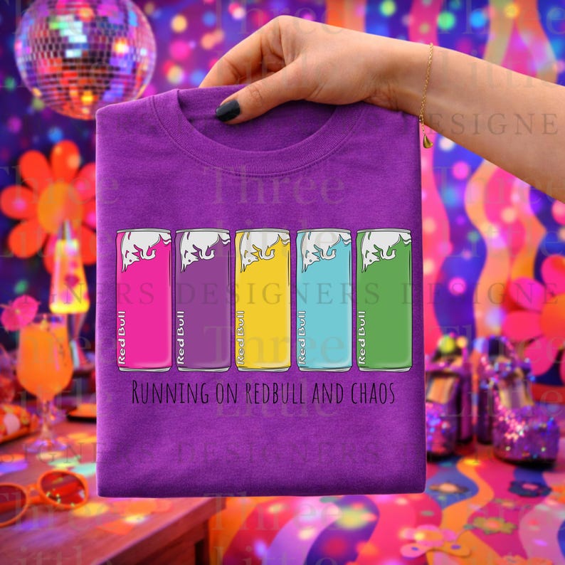 May include: Purple t-shirt featuring colorful Red Bull can graphics in pink, purple, yellow, blue, and green. The text "RUNNING ON REDBULL AND CHAOS" is printed below the cans. A hand holds the shirt.
