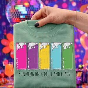 May include: Sage green t-shirt featuring a graphic of five Red Bull cans in pink, purple, yellow, blue, and green. The text "RUNNING ON REDBULL AND CHAOS" is printed below the cans. The shirt is held by a hand.