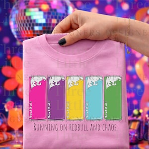 May include: Pink crew neck sweatshirt with a graphic of five colorful Red Bull cans. The cans are in shades of pink, purple, yellow, blue, and green. The text "RUNNING ON REDBULL AND CHAOS" is printed below the cans.