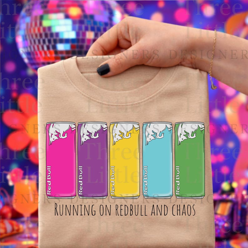 May include: Beige crew neck t-shirt featuring a graphic of five Red Bull cans in pink, purple, yellow, blue, and green. The text "RUNNING ON REDBULL AND CHAOS" is printed below the cans. The shirt is held up by a hand.