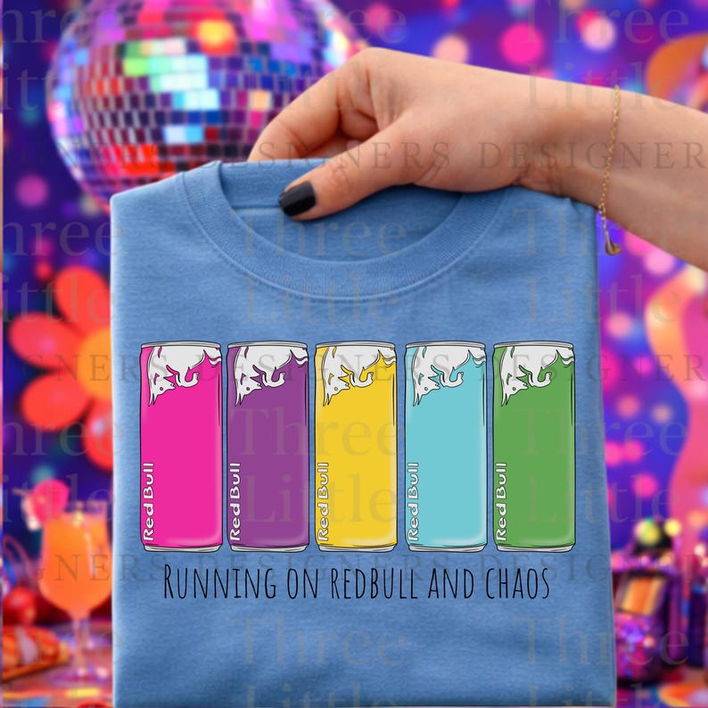 May include: A light blue t-shirt featuring five Red Bull energy drink cans in pink, purple, yellow, teal, and green. The text "RUNNING ON REDBULL AND CHAOS" is printed below the cans. The shirt is held up by a hand.
