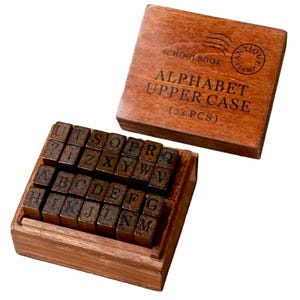 May include: Wooden box with 28 alphabet stamps in uppercase. The box is brown and has the text "SCHOOLBOOK" and "ALPHABET UPPER CASE (28 PCS)" on the top.