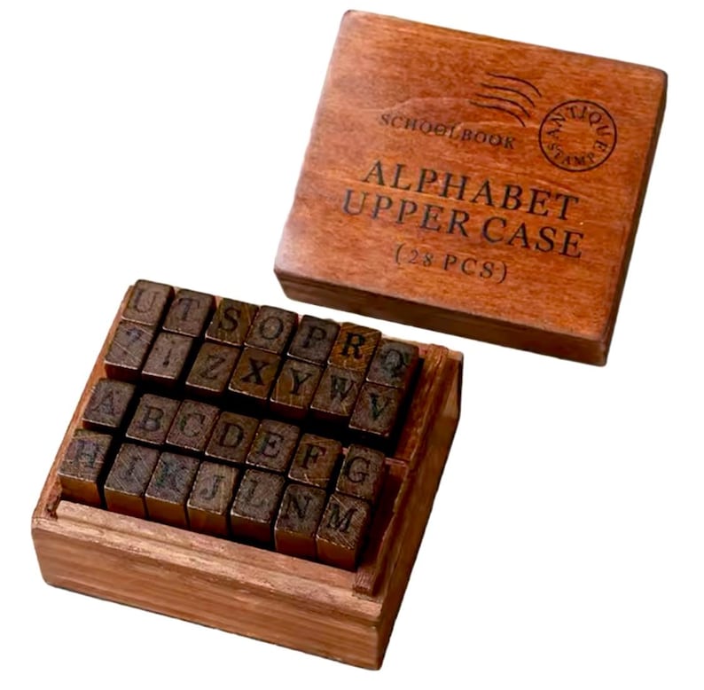 May include: Wooden box with 28 alphabet stamps in uppercase. The box is brown and has the text "SCHOOLBOOK" and "ALPHABET UPPER CASE (28 PCS)" on the top.