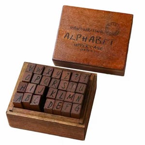 May include: A wooden box filled with 26 individual wooden alphabet stamps, each with a handwritten uppercase letter. The box has a brown lid with the text "HANDWRITING ALPHABET UPPERCASE (26 PCS)" on it.