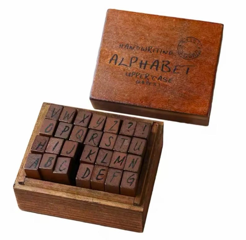 May include: A wooden box filled with 26 individual wooden alphabet stamps, each with a handwritten uppercase letter. The box has a brown lid with the text "HANDWRITING ALPHABET UPPERCASE (26 PCS)" on it.