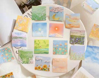 May include: A collection of square sticky notes with various landscape paintings. The notes feature scenes of fields, sunsets, mountains, and flowers in pastel colors. The notes are arranged on a white plate, with some scattered around.