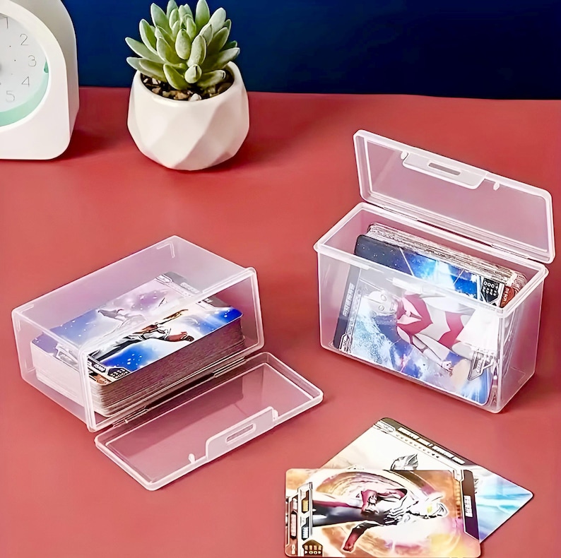 Puede incluir: Dos cajas de almacenamiento de pl&aacute;stico transparente con tapas, cada una llena de una pila de cartas coleccionables de colores. Las cajas est&aacute;n sobre una superficie rosa.