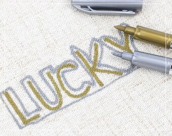 May include: Two metallic pens, one gold and one silver, are used to write the word "LUCKY" on a beige fabric surface. The word is outlined in silver and filled in with gold.