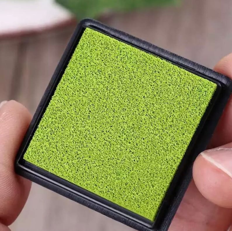 May include: A close-up of a green ink pad with a black plastic frame. The pad is made of a porous material and is designed for stamping.