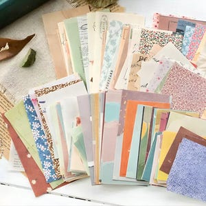 Op de afbeelding: Een verzameling diverse vintage papieren ephemera, waaronder ansichtkaarten, papieren met patronen en journaalkaarten. De papieren hebben een verscheidenheid aan kleuren, patronen en texturen, met bloemen en abstracte ontwerpen. Sommige papieren hebben tekst en de algehele esthetiek is vintage en artistiek.