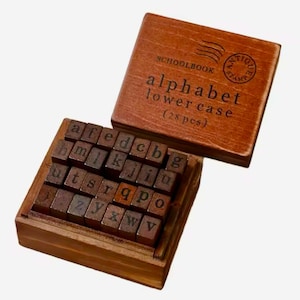 May include: Wooden box filled with 28 lowercase alphabet letter stamps. The box has a label that reads "Schoolbook alphabet lower case (28 pcs)" and a vintage stamp that says "Antique Stamp".