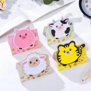Op de afbeelding: Vier kleurrijke dier-vormige sticky note houders. De houders zijn gevormd als een varken, koe, schaap en tijger. Elke houder heeft een schattig cartoon gezicht en staat op een klein kaartje met een herfstblad ontwerp.