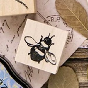 Può includere: Un timbro in gomma nero e bianco con un design di ape. L'ape ha strisce nere e gialle ed è in posizione di volo con le ali spiegate.