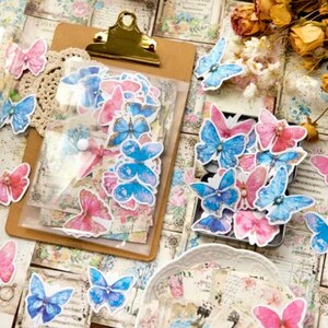 May include: A sheet of colorful butterfly stickers with blue and pink butterflies. The stickers are arranged in a clear plastic bag and on a brown clipboard. The stickers are perfect for scrapbooking, journaling, and other craft projects.