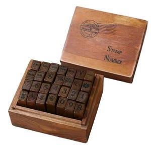 May include: A wooden box with a set of 30 alphabet and number stamps. The box is brown and has a label that says "Stamp Number". The stamps are arranged in rows and are made of wood.