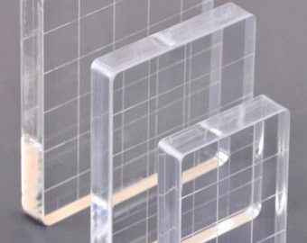 May include: Three clear acrylic stamp blocks of different sizes. The blocks have a grid pattern etched into them.