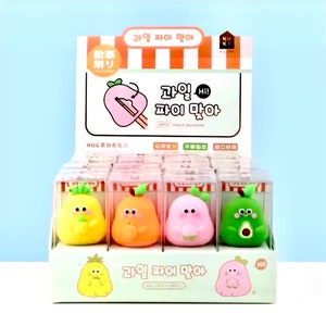 May include: A display of 24 colourful fruit-shaped pencil sharpeners. The sharpeners are in individual clear plastic boxes. The boxes are arranged in rows on a green and white display stand. The display stand has the text "과일 파이 맞아" in Korean.