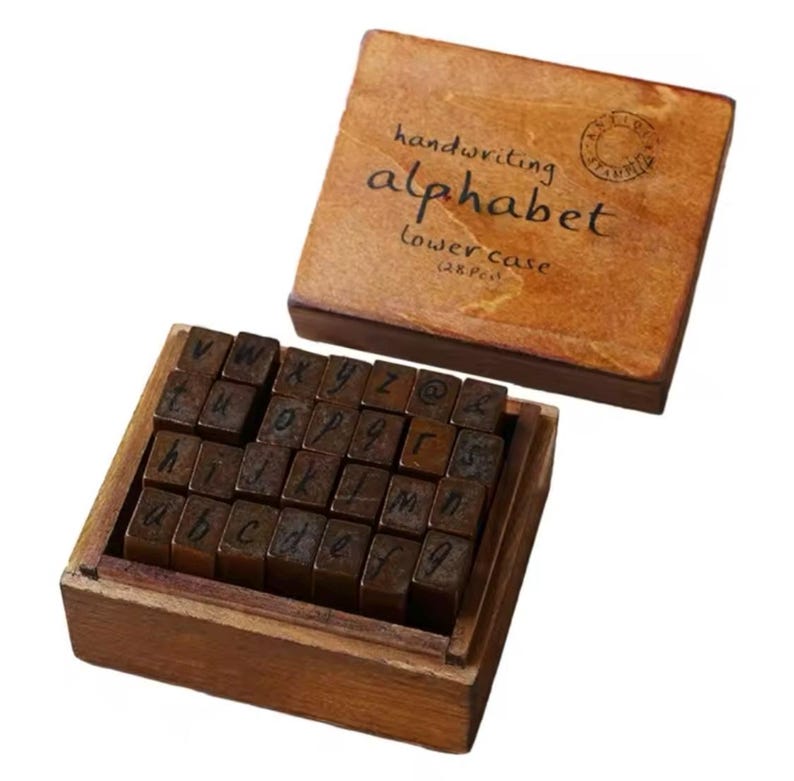 May include: A wooden box filled with individual wooden stamps of the lowercase alphabet, numbers, and symbols. The box lid is brown and has the text "handwriting alphabet lowercase (28 pcs)" printed on it.