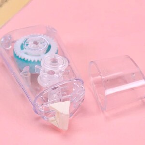 May include: A clear plastic tape dispenser with a blue and white gear mechanism. The dispenser has a white triangular tape cutter. The dispenser is open and the tape is visible.