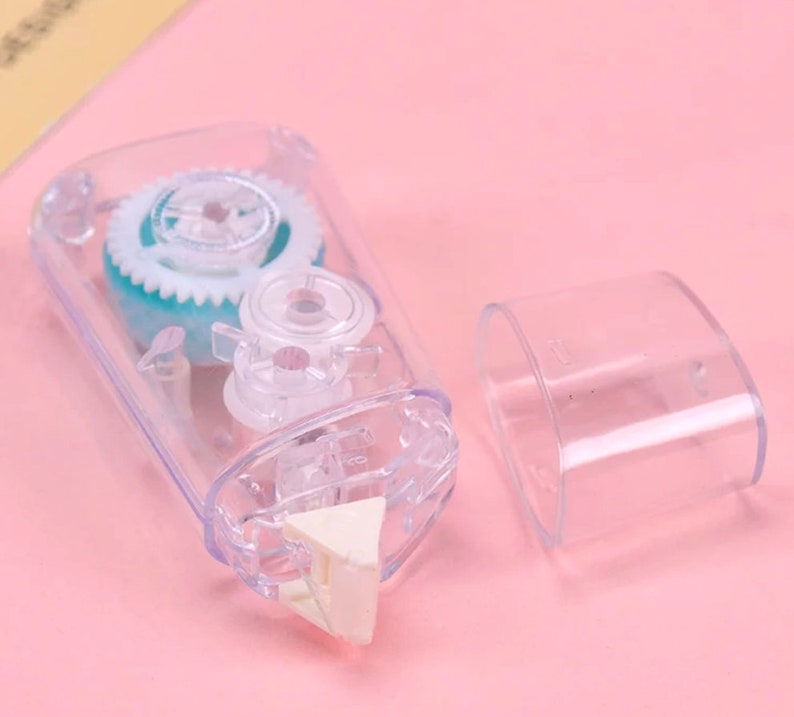 May include: A clear plastic tape dispenser with a blue and white gear mechanism. The dispenser has a white triangular tape cutter. The dispenser is open and the tape is visible.