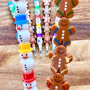 Pode incluir: Um conjunto de lápis coloridos com tema natalino com toppers de boneco de neve e homem de gengibre. Os lápis estão empilhados em uma torre com os toppers virados para a câmera.