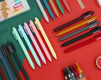 Puede incluir: Una variedad de bolígrafos de gel de colores, incluyendo rosa pastel, azul, verde y amarillo, y colores oscuros como negro, marrón, gris y rojo. Los bolígrafos están dispuestos sobre una superficie roja y verde.