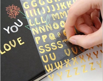 May include: Gold alphabet letter stickers on a clear sheet. The letters are arranged in alphabetical order and are ready to be peeled off and used for crafting projects.