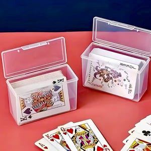 Puede incluir: Dos cajas de pl&aacute;stico transparente con tapas, cada una contiene una baraja de cartas para jugar. Una caja tiene una baraja de cartas est&aacute;ndar con un rey de picas en la parte superior. La otra caja tiene una baraja de cartas de comod&iacute;n con un comod&iacute;n en la parte superior. Las cajas est&aacute;n sobre una superficie roja.