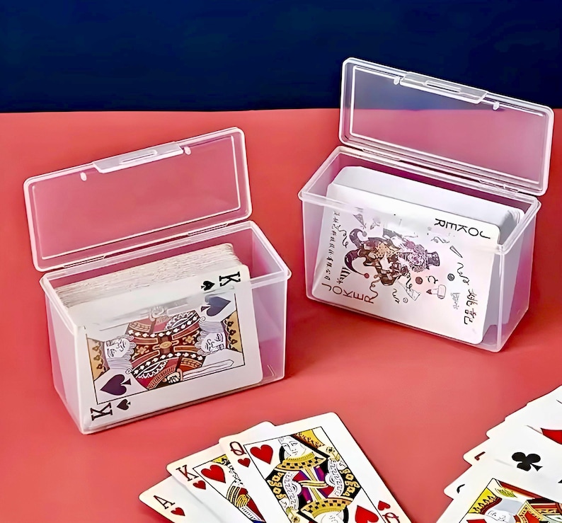 Puede incluir: Dos cajas de pl&aacute;stico transparente con tapas, cada una contiene una baraja de cartas para jugar. Una caja tiene una baraja de cartas est&aacute;ndar con un rey de picas en la parte superior. La otra caja tiene una baraja de cartas de comod&iacute;n con un comod&iacute;n en la parte superior. Las cajas est&aacute;n sobre una superficie roja.
