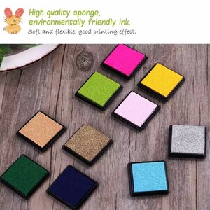 May include: Ten ink pads in various colors, including green, pink, blue, gold, and silver. The ink pads are square and have a black plastic frame. The ink pads are arranged in a circle on a wooden surface.