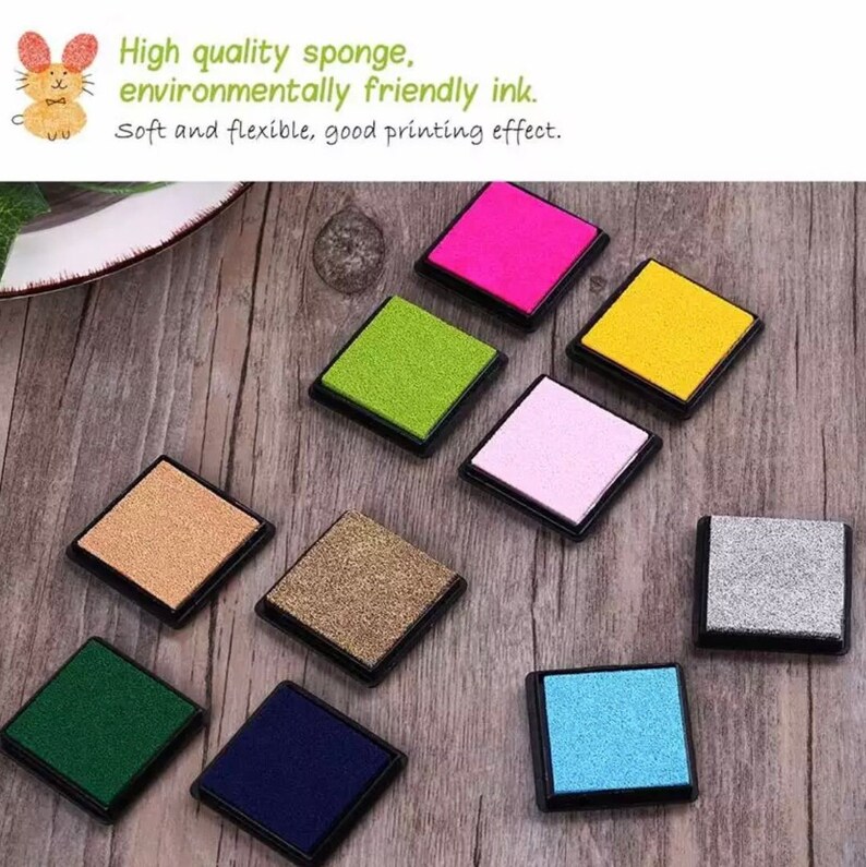 May include: Ten ink pads in various colors, including green, pink, blue, gold, and silver. The ink pads are square and have a black plastic frame. The ink pads are arranged in a circle on a wooden surface.