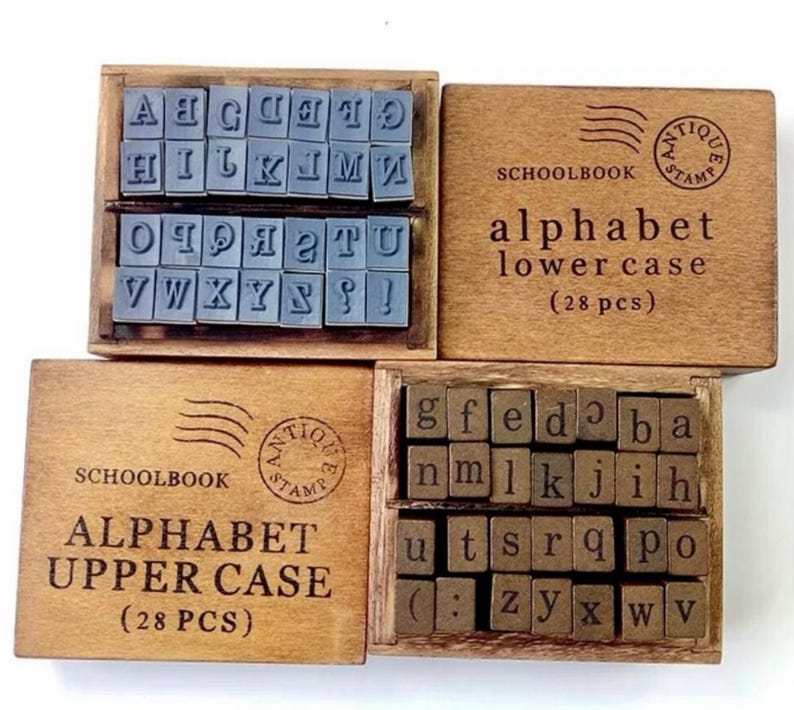 May include: Two wooden boxes containing alphabet stamps. The top box contains uppercase letters and the bottom box contains lowercase letters. Each box has a label that says "Schoolbook" and "Antique Stamp".