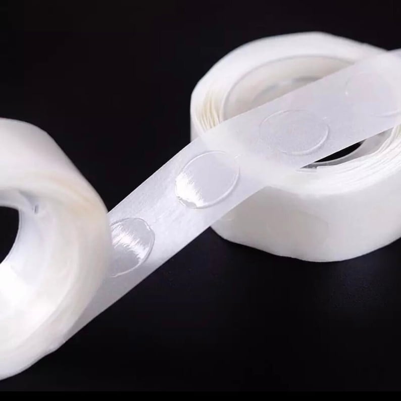 May include: A roll of clear adhesive dots on a white tape dispenser. The dots are designed to attach balloons to walls or ceilings.