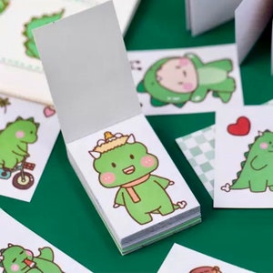 以下が含まれることがあります： かわいい緑色の恐竜のイラストが描かれた、小さな白いメモ帳の束。それぞれのメモ帳には、自転車に乗った恐竜、マフラーを巻いた恐竜、ハートを持った恐竜など、異なる恐竜のデザインが描かれています。