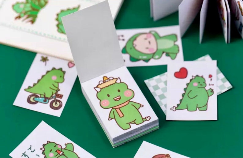 以下が含まれることがあります： かわいい緑色の恐竜のイラストが描かれた、小さな白いメモ帳の束。それぞれのメモ帳には、自転車に乗った恐竜、マフラーを巻いた恐竜、ハートを持った恐竜など、異なる恐竜のデザインが描かれています。